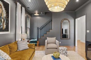 A living room with a sofa, armchair, and ottoman, a staircase, and a large mirror. - Painted Lady - Pool, Hot Tub & Walk to Main (Fredericksburg)