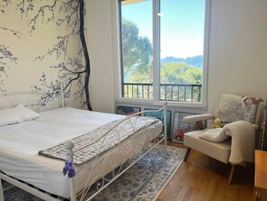 Un dormitorio con cama, una silla, un ventanal con vistas y un diseño de árbol en la pared.