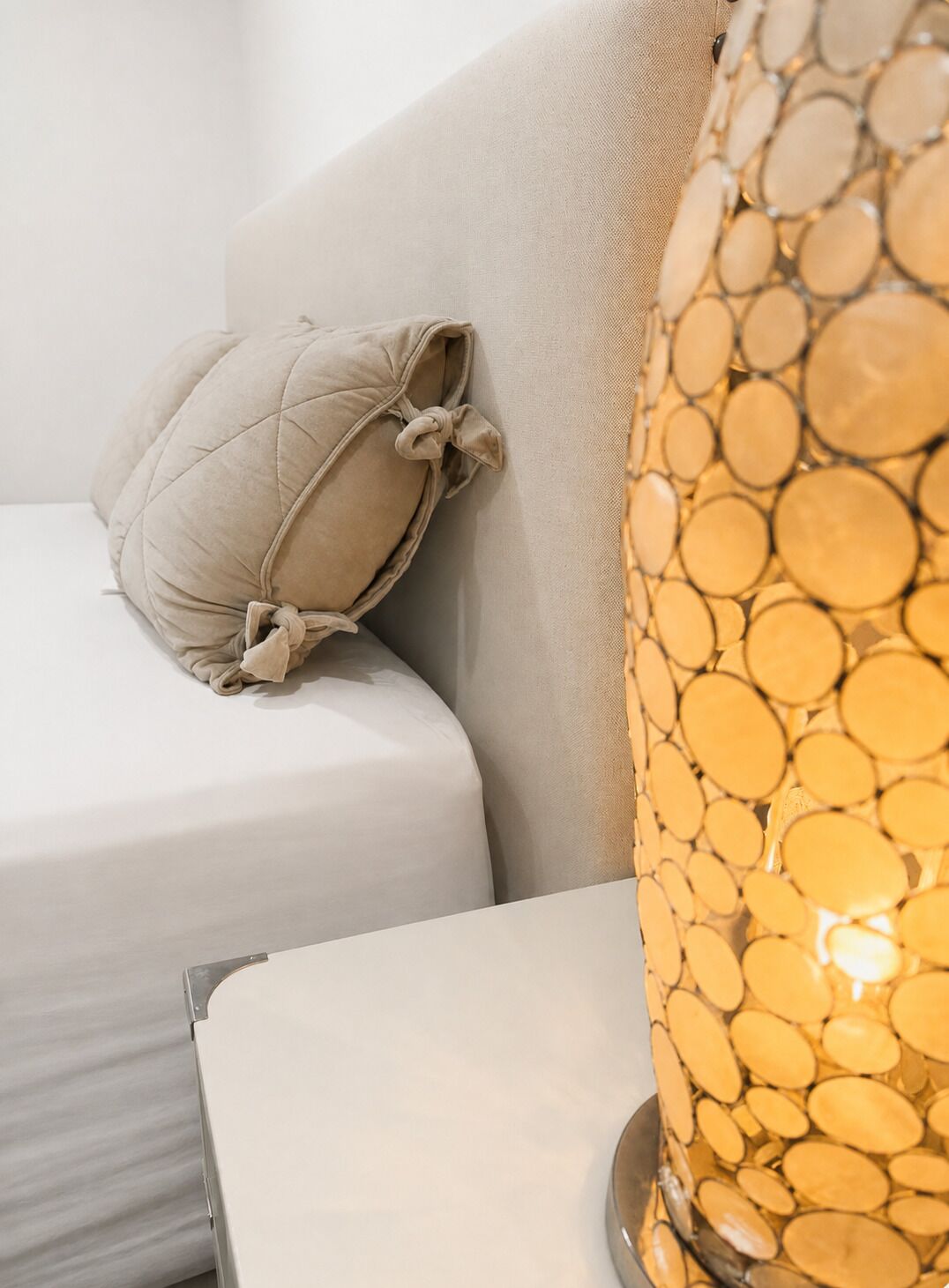 Ein Bett mit weißer Matratze und beigem Kopfkissen, ein Nachttisch und eine Lampe mit einer gläsernen Leuchte mit Struktur.