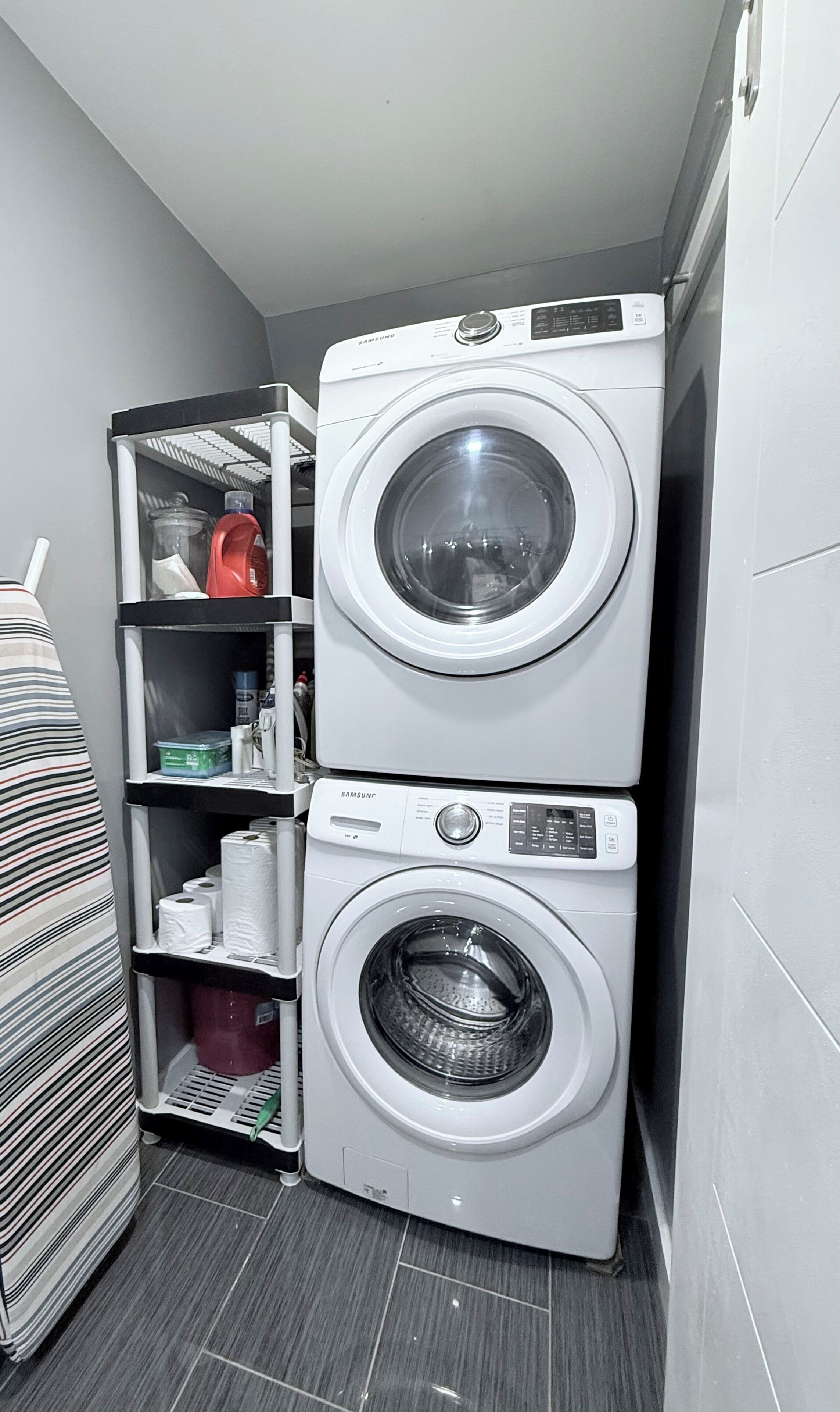 A compact laundry area with two stacked washing machines, a shelf with various items, and a striped fabric roll.