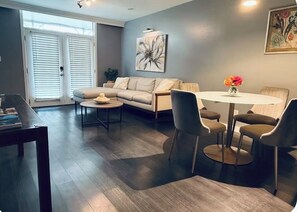 A modern living room with a sofa, coffee table, and chairs. There is a dining table with chairs and a vase of flowers. A painting is on the wall, and there are windows with blinds.