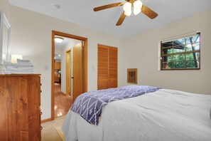 Une chambre à coucher comprenant un lit, une commode en bois, un ventilateur de plafond, une fenêtre donnant sur des arbres et une porte menant à la salle de bain.
