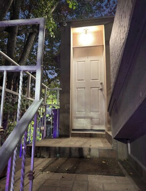 Un conjunto de escalones exteriores que conducen a una puerta blanca con una luz encima, rodeado de árboles y una barandilla de metal.