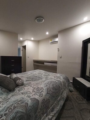 A bedroom with a bed, dresser, mirror, and built-in air conditioning unit.
