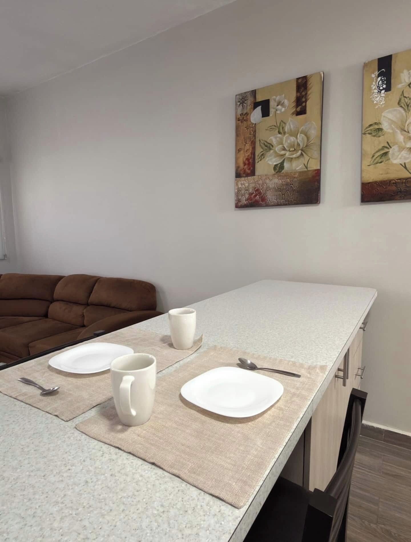A dining area with a table set for two, two white plates, two white cups, and a spoon. There is a brown sofa in the background.