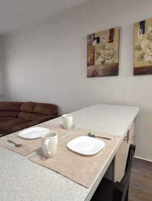 A dining area with a table set for two, two white plates, two white cups, and a spoon. There is a brown sofa in the background.