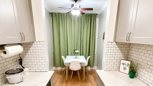A compact kitchen with white cabinets, a dining table with chairs, a ceiling fan, and a green curtain. - Charming Downtown Studio - Walk to Toyota Center (Houston)