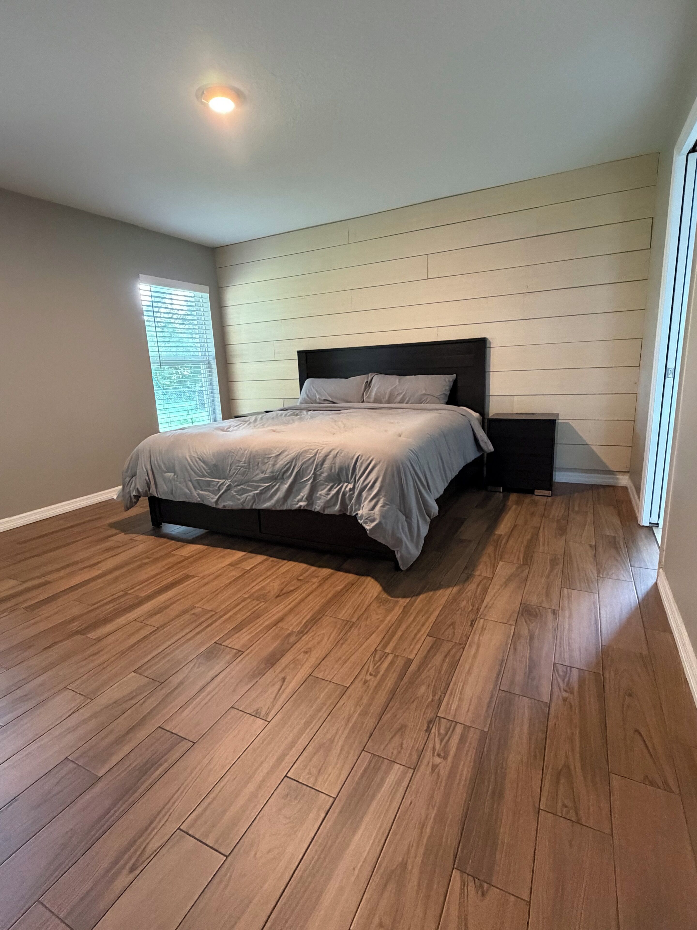 A bedroom with a bed, wooden flooring, a window with blinds, and a nightstand.