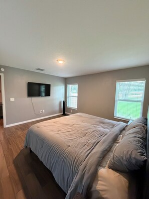 A bedroom with a large bed, a flat-screen TV mounted on the wall, and two windows with blinds. - 3-bedroom house near Brownwood • Pet Friendly (Wildwood)