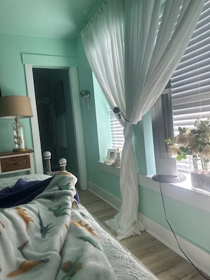 A bedroom with a bed, a nightstand, a lamp, a window with curtains, and a radiator. - Perfect 4-bedroom house with AC in quaint Aransas Pass (Aransas Pass)