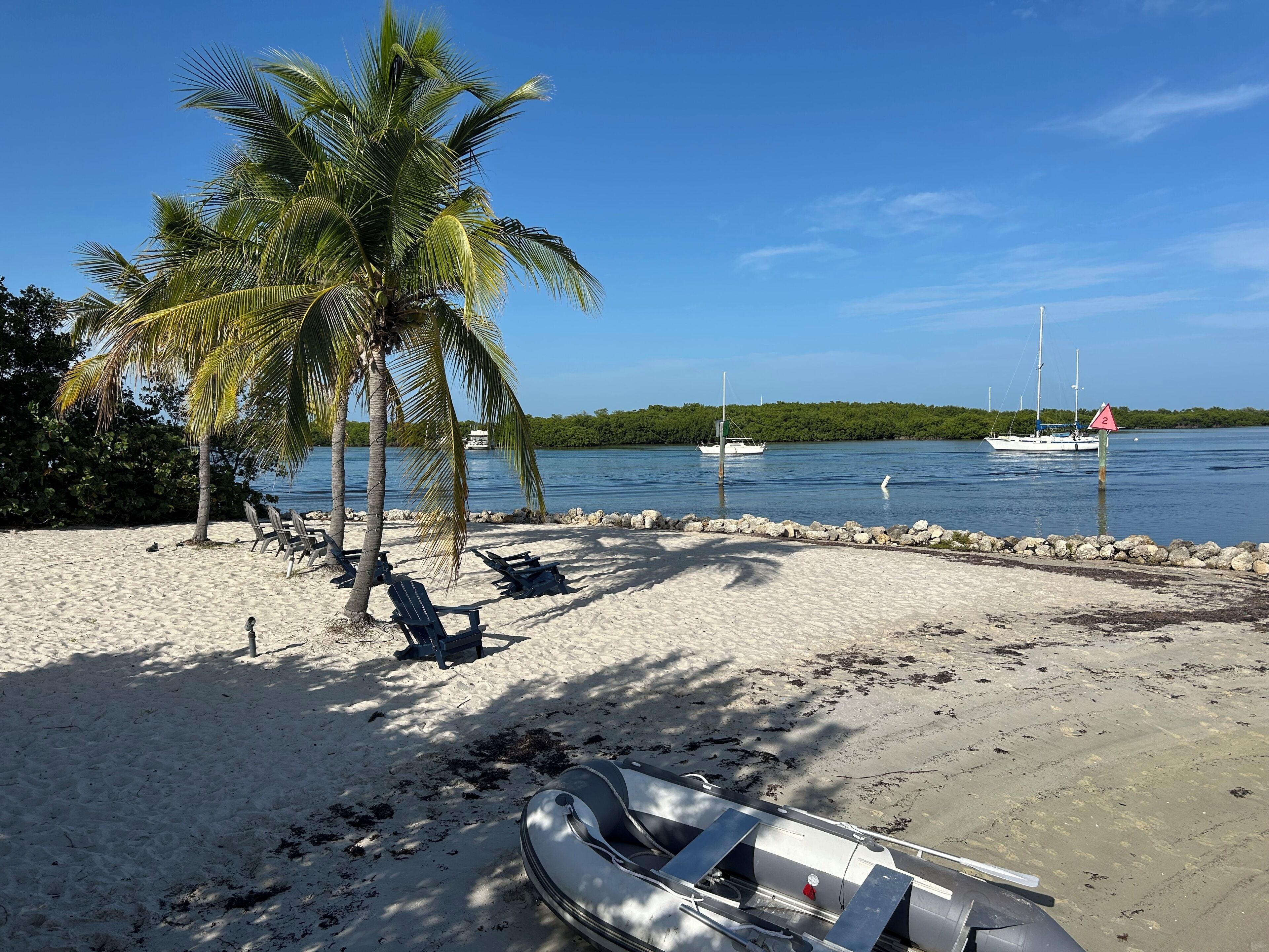Una playa con palmeras, sillas de descanso, un bote y un canal con barcos.