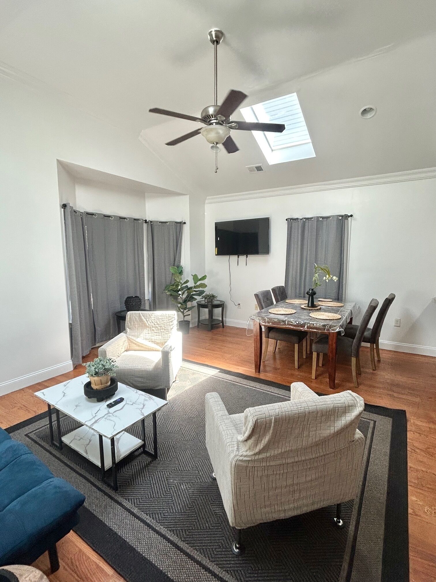 A spacious living area with a ceiling fan, a dining table with chairs, a TV mounted on the wall, and a large window with curtains.