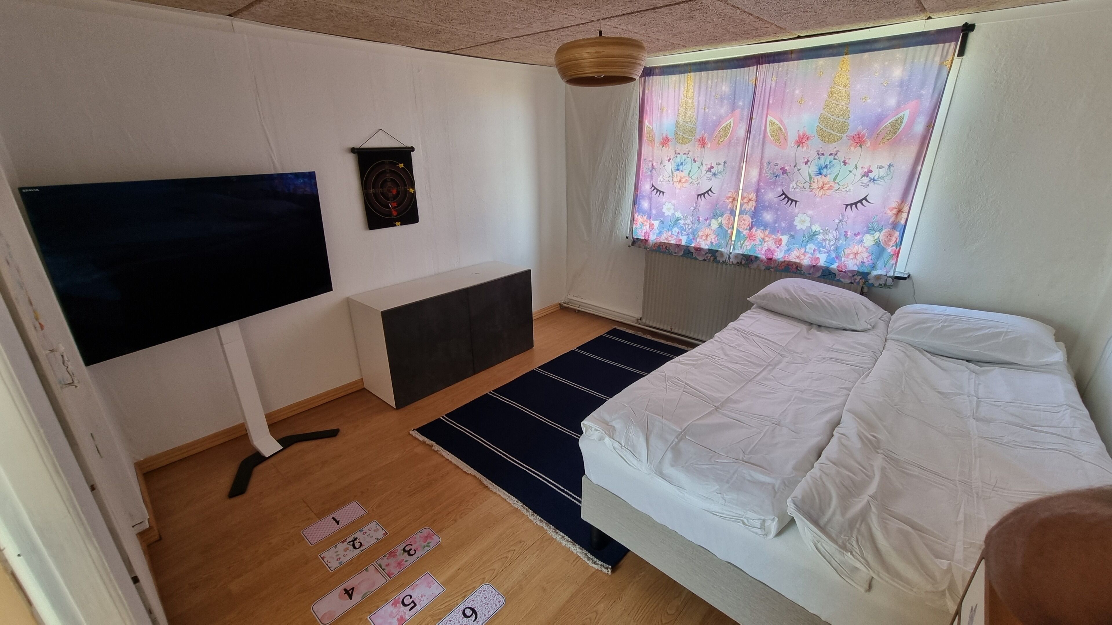 Ein Schlafzimmer mit Bett, Fernseher, einer Wandablage, einem Fenster mit Vorhängen und einem Holzboden mit nummerierten Matten.
