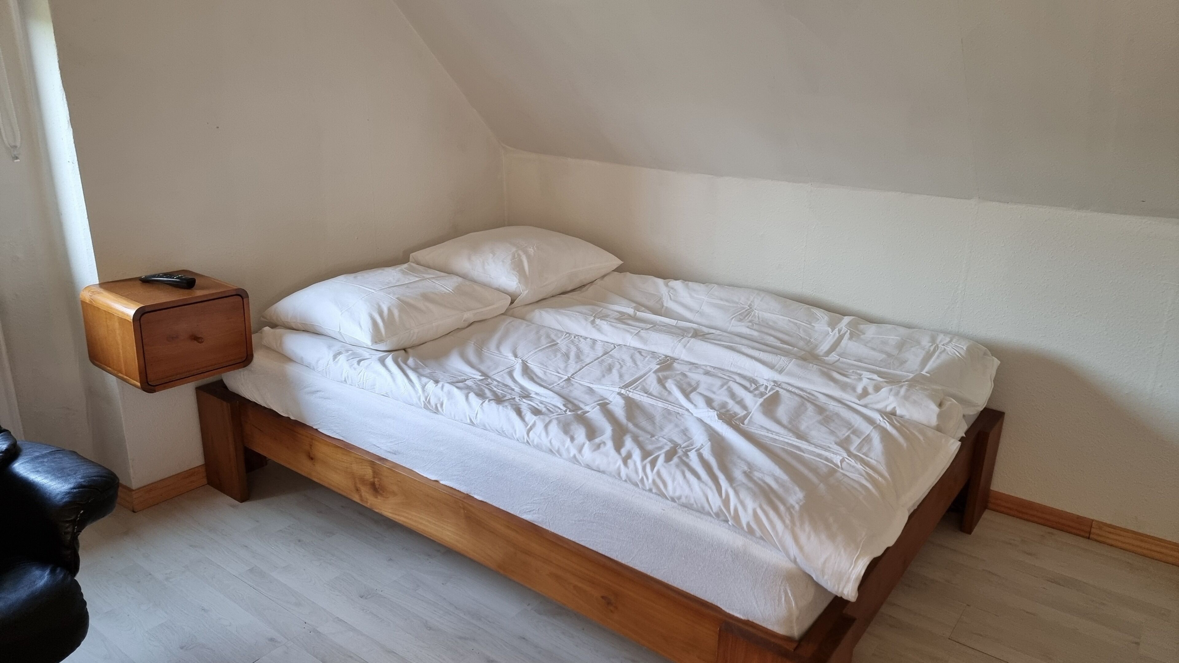 A single bed with a wooden frame and white bedding in a room with white walls and a sloped ceiling.