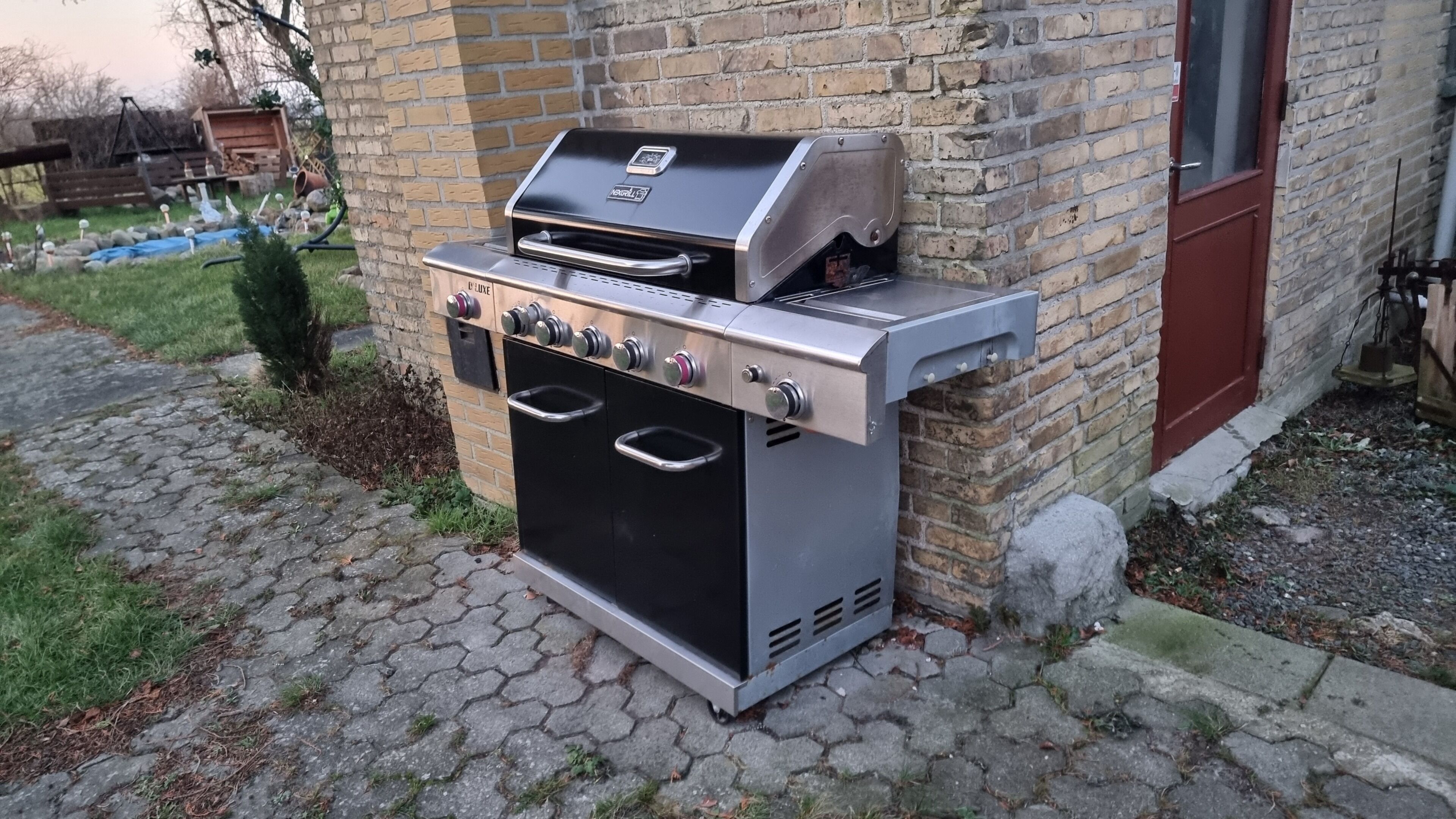 An outdoor gas grill with four burners, a built-in oven, and a digital control panel, placed on a paved patio next to a brick wall.