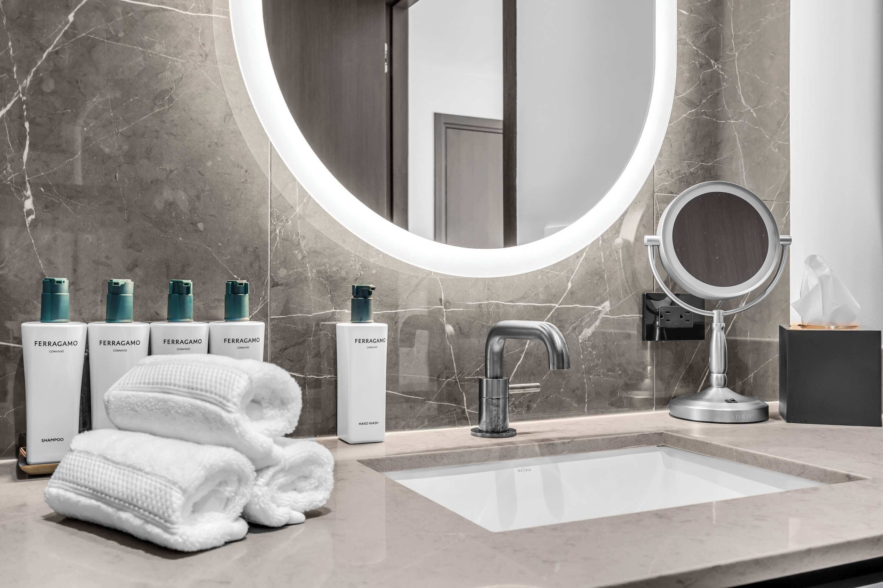 A bathroom with a marble sink, a round mirror, and toiletries.