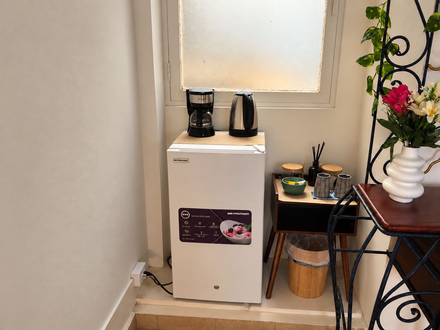 Uma geladeira compacta com visor digital, uma cafeteira e uma mesa pequena com um vaso de flores.