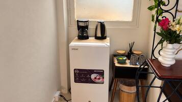 Uma geladeira compacta com visor digital, uma cafeteira e uma mesa pequena com um vaso de flores.