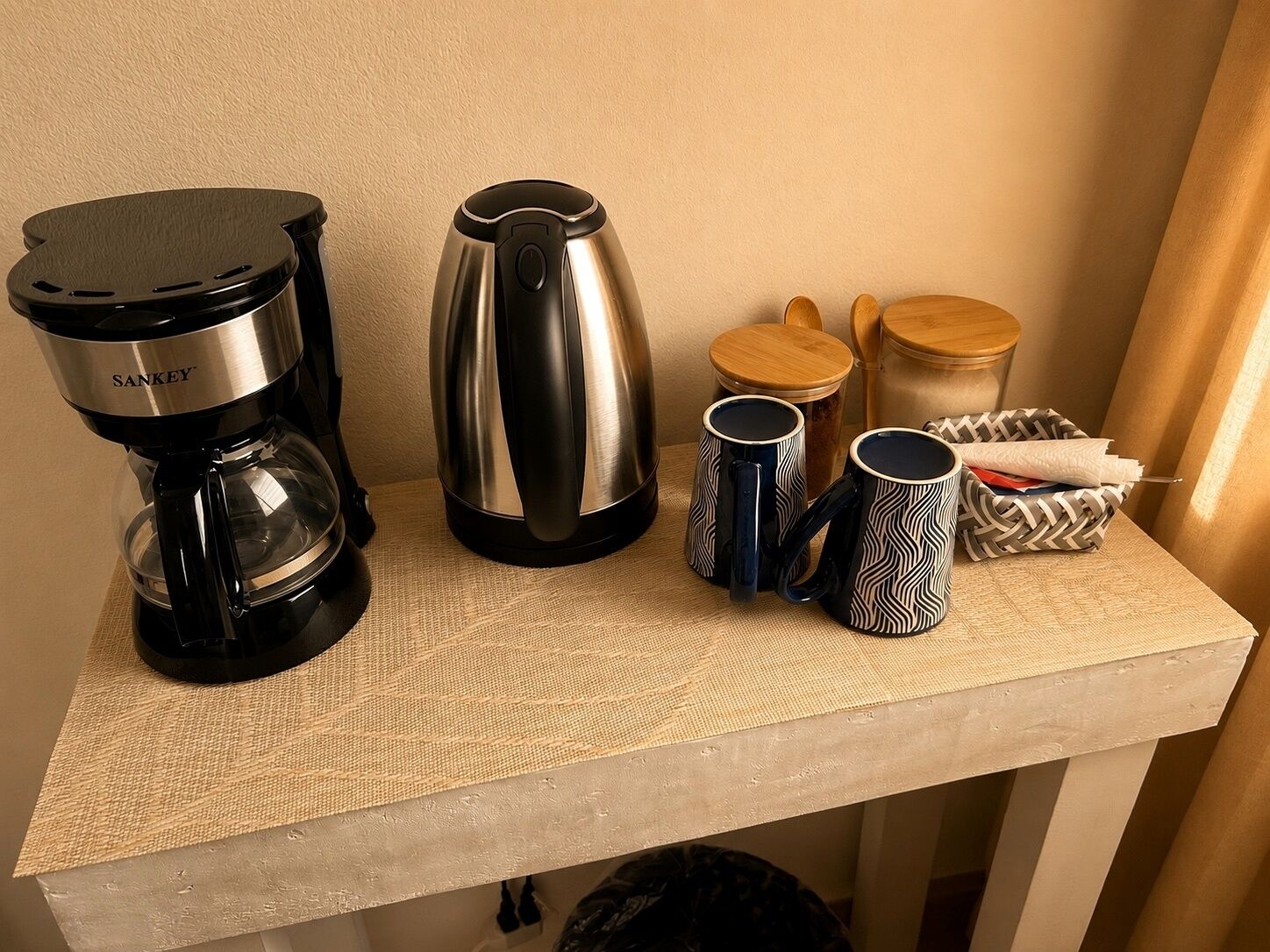 A kitchenette with a coffee maker, a kettle, and two mugs on a textured table.