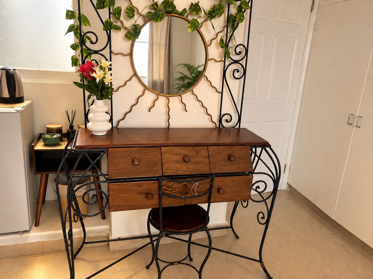 Une coiffeuse en bois avec un miroir, un petit tabouret et un vase rempli de fleurs.