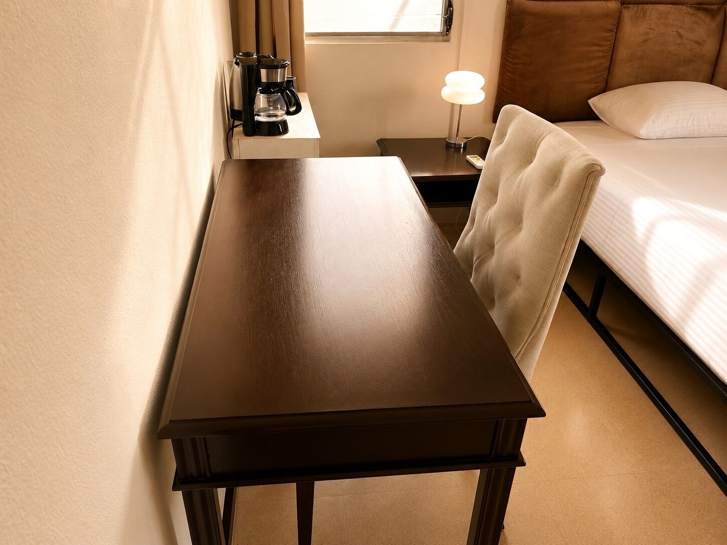 Ein Hotelzimmer mit einem Holztisch, einem Bett mit weißer Bettwäsche, einem Nachttisch mit Lampe und einem Fenster mit Vorhängen.