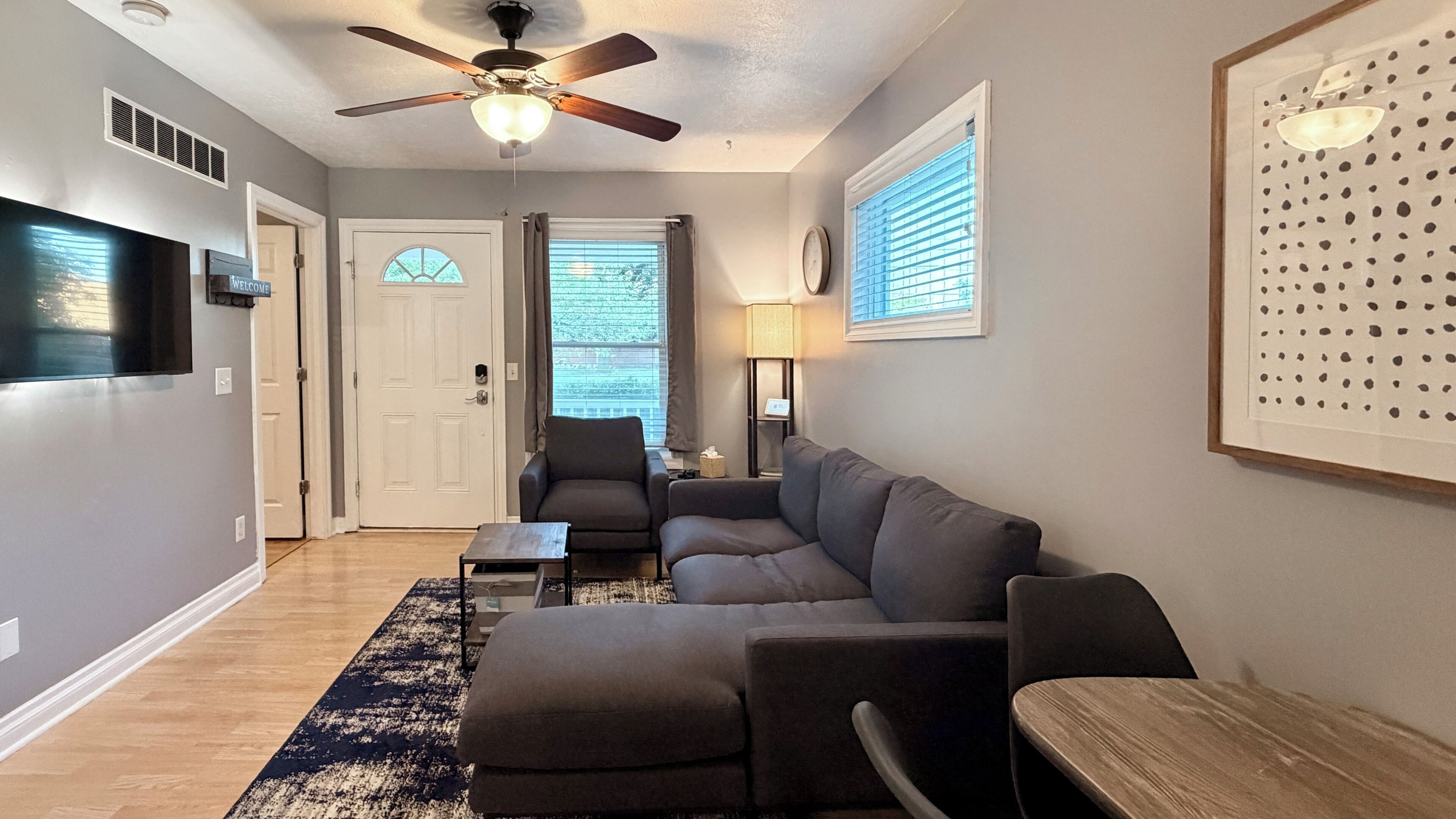 A living room with a sectional sofa, a coffee table, a TV, a ceiling fan, and a framed artwork on the wall.