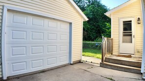 A residential garage with a white door, a wooden front door, and a small porch with a wooden step. - The Comfort Street House - Pet-Friendly Home Near Downtown Lansing w/Office (Lansing)
