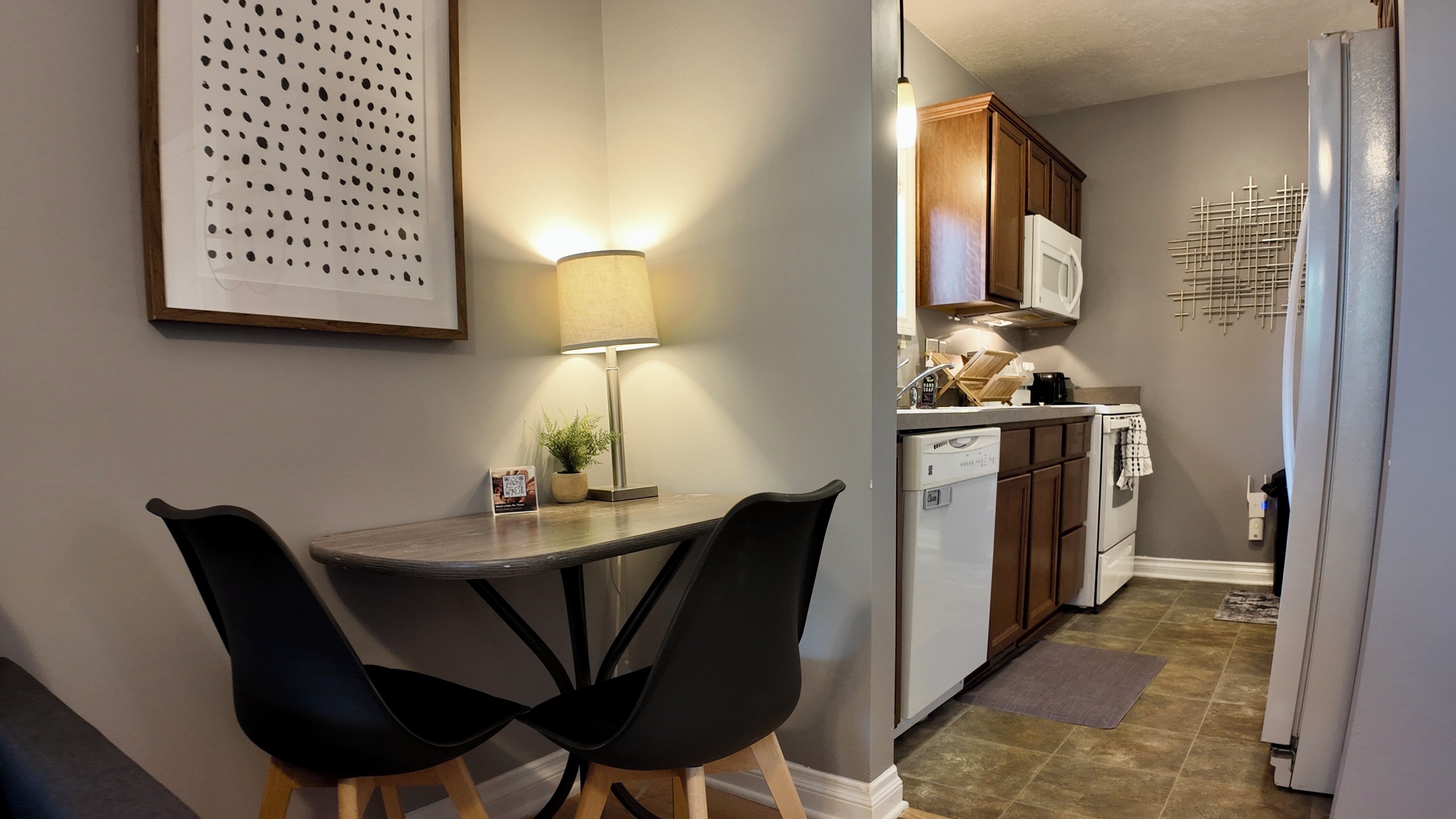 A compact kitchen with a dining area, featuring a table with two chairs, a lamp, and a refrigerator.