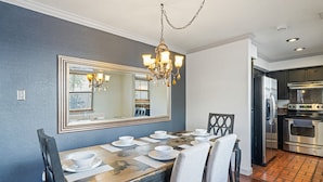 Un coin repas agrémenté d’un grand miroir, d’un lustre, d’une table en bois dressée avec de l’assiette et des bols blancs, et d’une cuisine équipée d’appareils électroménagers en acier inoxydable.