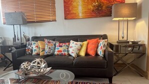 Sala de estar com sofá preto, almofadas coloridas, mesa de centro de vidro, luminária e um quadro na parede.