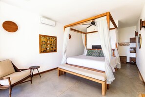 Ein Himmelbett mit Deckenventilator, ein Holzstuhl, ein kleiner runder Tisch und eine Wand-Klimaanlage.