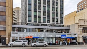 Ein mehrstöckiges Gebäude mit moderner Fassade, ein Restaurant mit sichtbarer Speisekarte und ein Parkplatz mit parkenden Autos.