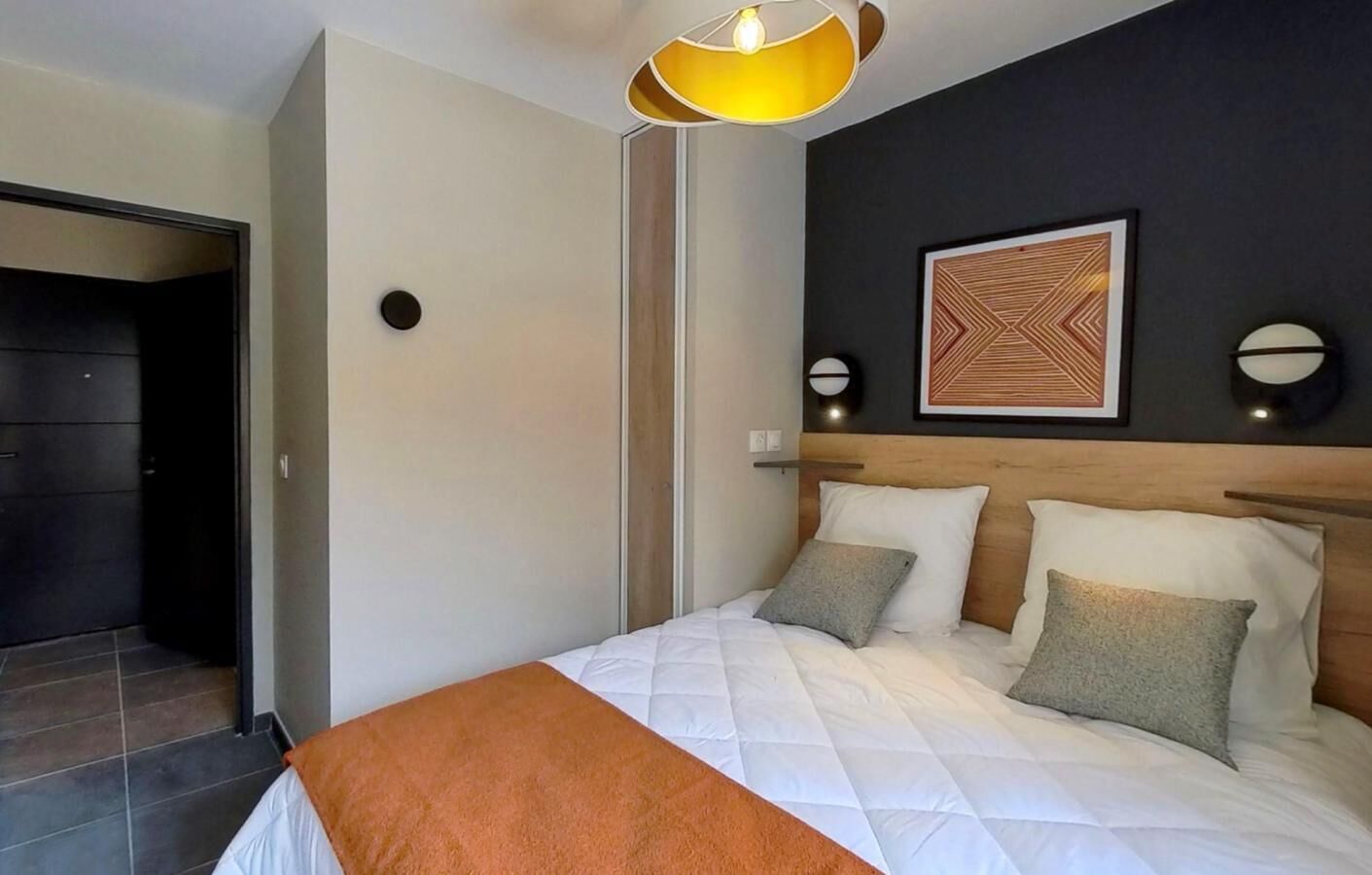 Una camera d'albergo con un letto, una testiera, un'opera d'arte appesa al muro e una porta che dà su un'altra stanza.