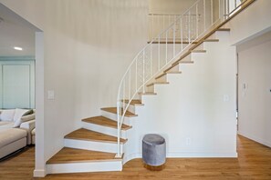 Una escalera con escalones de madera y barandillas blancas, un otoman gris redondo y una pared de color claro.