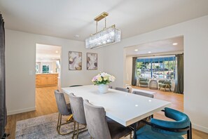 Un comedor moderno con una mesa blanca, sillas grises y azules, una araña y un jarrón de flores.