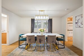 Un coin repas avec une table blanche, quatre chaises, un lustre et une fenêtre avec des rideaux.