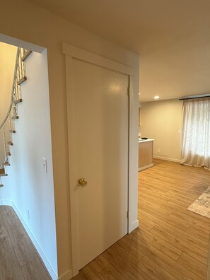 Ein Zimmer mit Holzboden, einer weißen Tür, einer Treppe und einem Fenster mit Vorhängen.