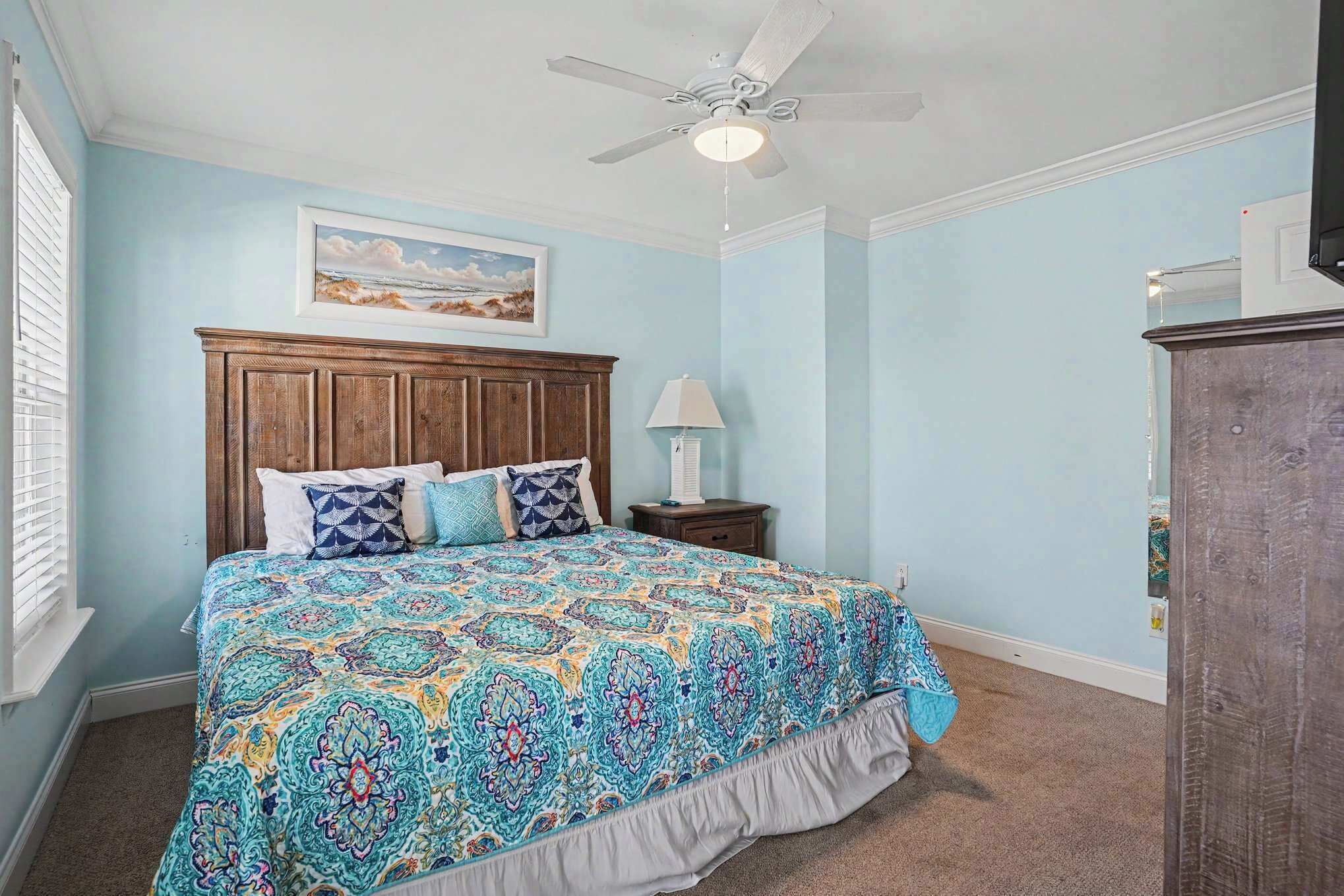 A bedroom with a bed, a nightstand, a lamp, a ceiling fan, and a painting on the wall.