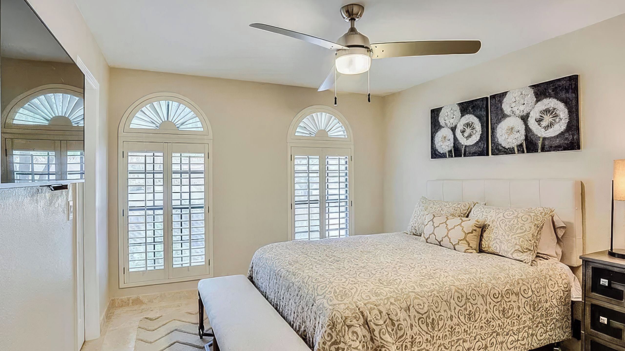 A bedroom with a bed, a ceiling fan, a window with shutters, and a painting on the wall.