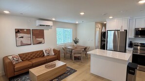 Een moderne keuken met roestvrijstalen apparatuur, een eethoek met een ronde tafel en stoelen, en een woonkamer met een bruine bank en salontafel.