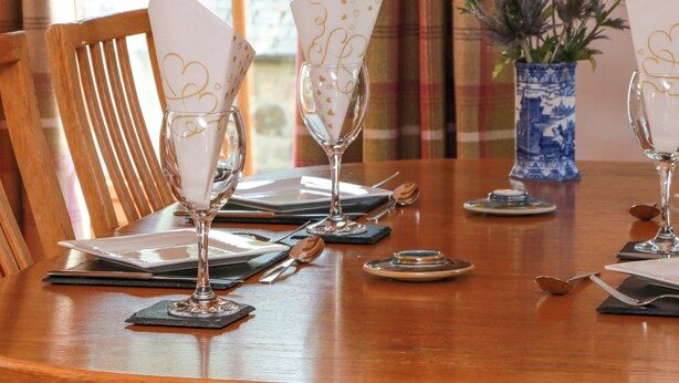 Una mesa de comedor dispuesta con copas de vino, platos y cubiertos, con un jarrón azul y cortinas al fondo.