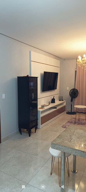 A modern living room with a flat-screen TV mounted on the wall, a dark wooden cabinet, a white console table, and a dining area with a marble table and chairs. - Beautiful apartment with ocean views on the Porto Seguro coastline, close to the best beaches. (Porto Seguro)