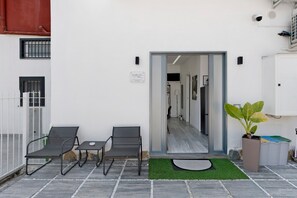 Ein Sitzbereich im Freien mit zwei Stühlen, einem Tisch, einem Blumentopf und einer Tür zu einem Innenraum.