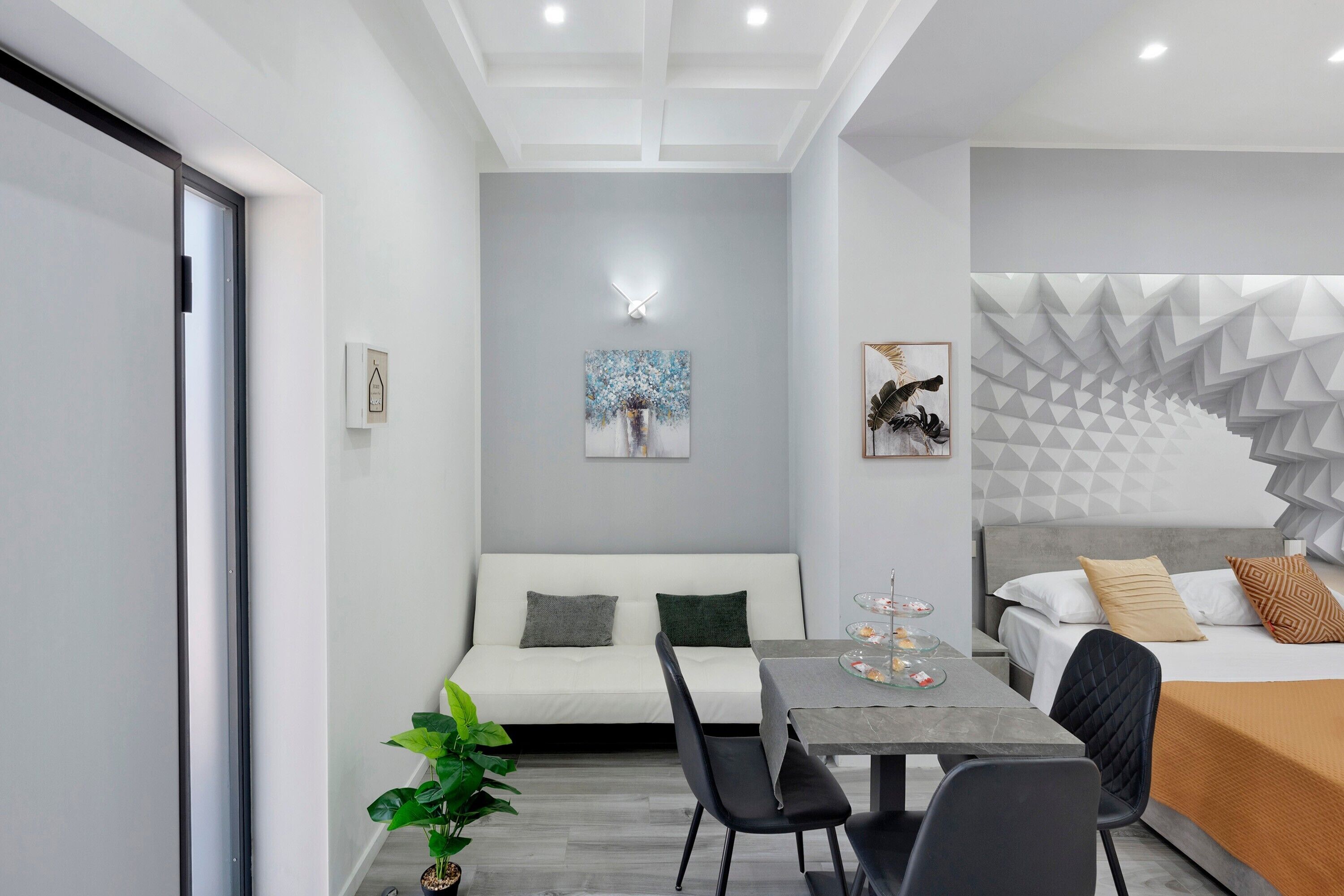 A modern hotel room with a sofa, dining table, chairs, a bed with a geometric headboard, and a wall-mounted artwork.