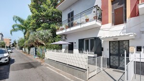 Une rue résidentielle avec un immeuble blanc doté d’un balcon orné de plantes en pot, d’une entrée fermée par un portail et d’un ciel dégagé.