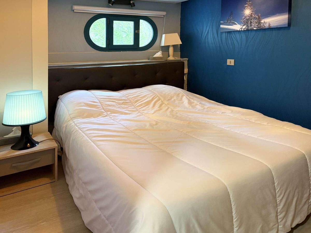 A neatly made bed in a room with a window, a bedside lamp, and a wall-mounted picture.