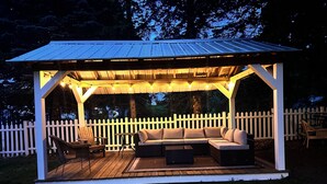 A covered outdoor seating area with a white picket fence, wooden deck, and string lights.