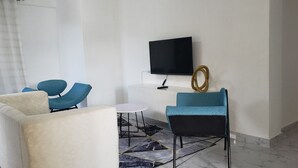 Ett modernt hotellrum med en vit soffa, två blå fåtöljer, ett litet runt bord, en platt-TV som är monterad på väggen och en matta med marmormönster.