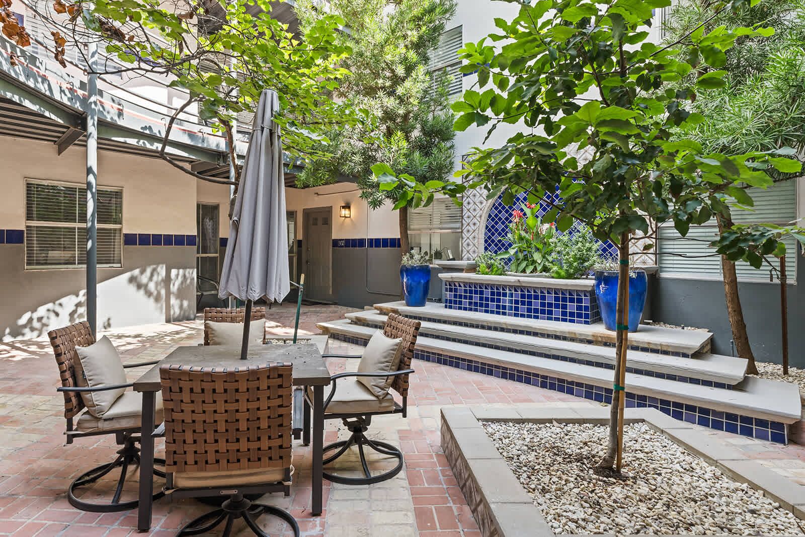 An outdoor patio with a dining table, chairs, and an umbrella. There are steps leading to a seating area with blue tiles and potted plants.