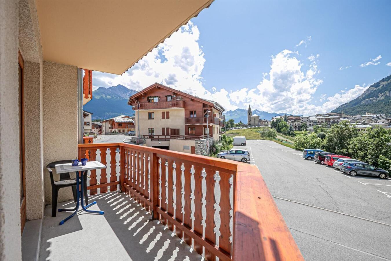 A balcony with a table and chairs, a view of a mountain, and a parking area with cars.
